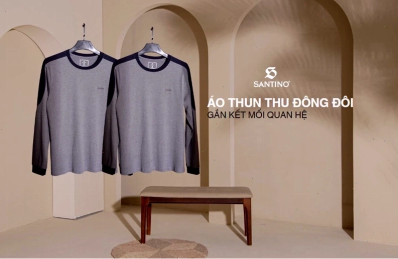 Cặp đôi mặc áo sweater đôi màu kem và nâu, ngồi cạnh nhau trên ghế đá công viên, trao nhau ánh nhìn trìu mến